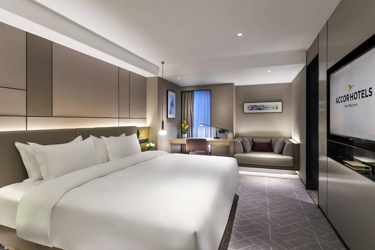 Een moderne hotelkamer met een groot bed, een bank, een bureau en een televisie waarop de naam van het hotel staat.