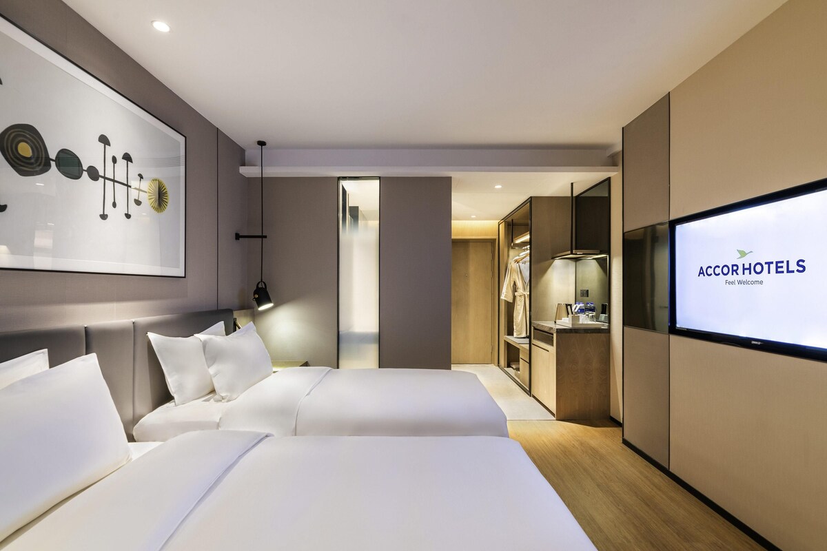 Ein modernes Hotelzimmer mit zwei Betten, einem großen Kunstwerk an der Wand, einem Fernseher mit der Anzeige 'AccorHotels' und einem begehbaren Kleiderschrank.