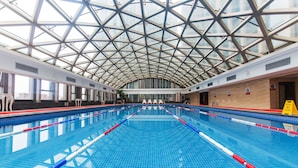 Une piscine intérieure avec des couloirs bien délimités, un plafond haut surmonté d’une verrière, et une vue sur le paysage urbain environnant.