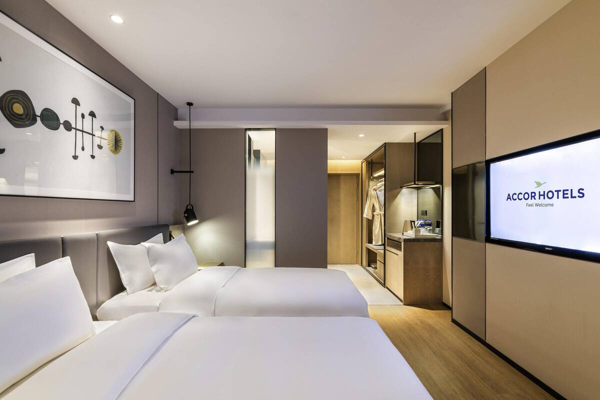 Een moderne hotelkamer met twee bedden, een groot kunstwerk aan de muur, een televisie waarop 'AccorHotels' staat en een inloopkast.