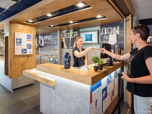 A hotel reception area with a wooden counter, a blue vase, and a potted plant. There are informational posters on the wall and a sign for ibis budget hotels. - ibis budget Manosque Cadarache (Manosque)