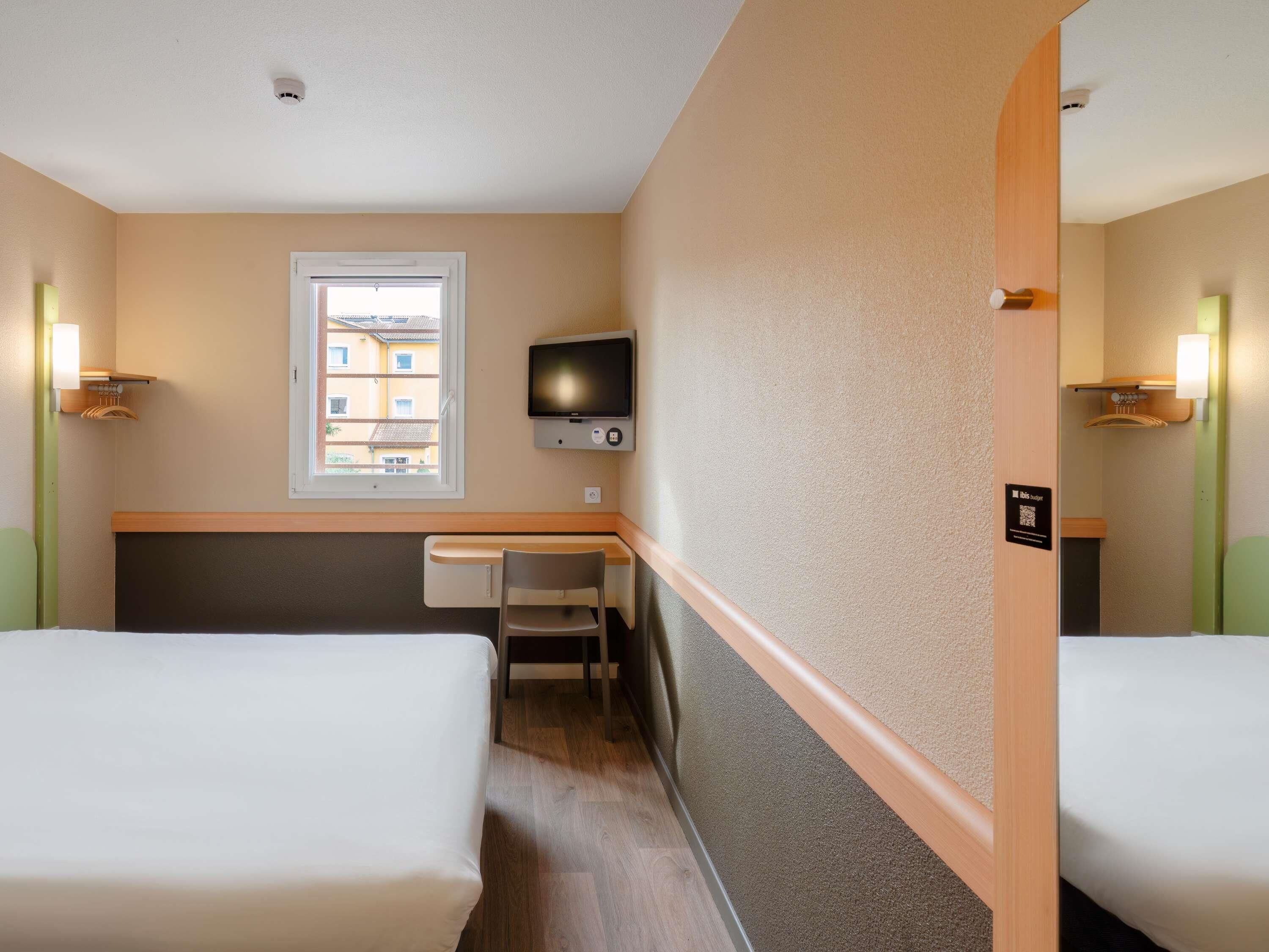 Habitación de hotel con dos camas, un escritorio, una silla, un televisor y una ventana con vista a edificios.