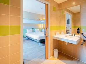 A hotel room with a bed, a bathroom with a sink and mirror, and a trash bin. - ibis budget Manosque Cadarache (Manosque)