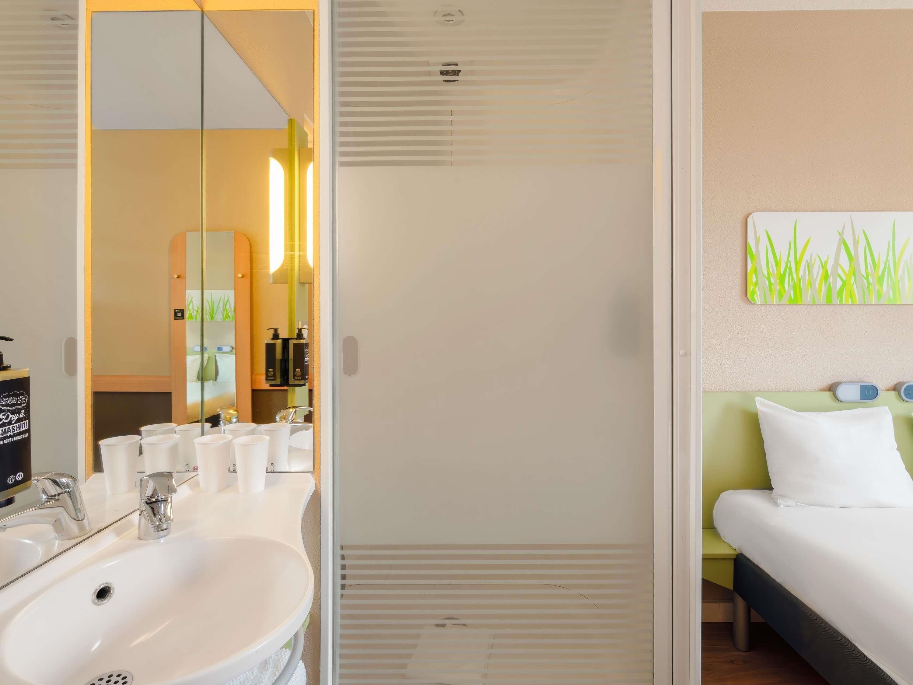 Un baño de hotel con lavamanos, espejo y artículos de aseo. Hay una cama con cabecera verde y almohada blanca.