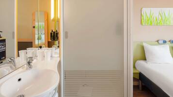 Un baño de hotel con lavamanos, espejo y artículos de aseo. Hay una cama con cabecera verde y almohada blanca.