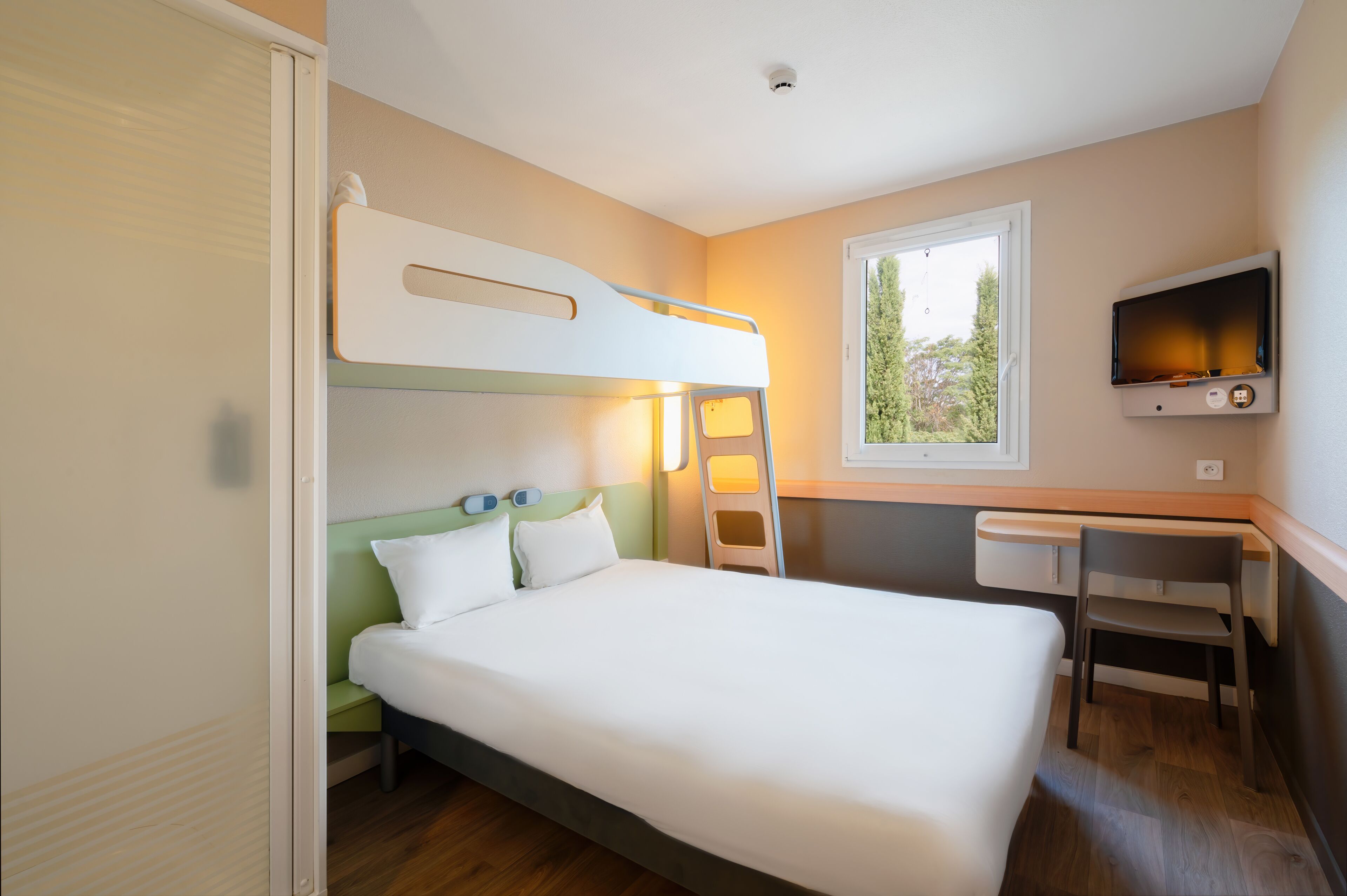 Una cama individual superpuesta con escritorio y silla, televisión y ventana con vista a árboles.