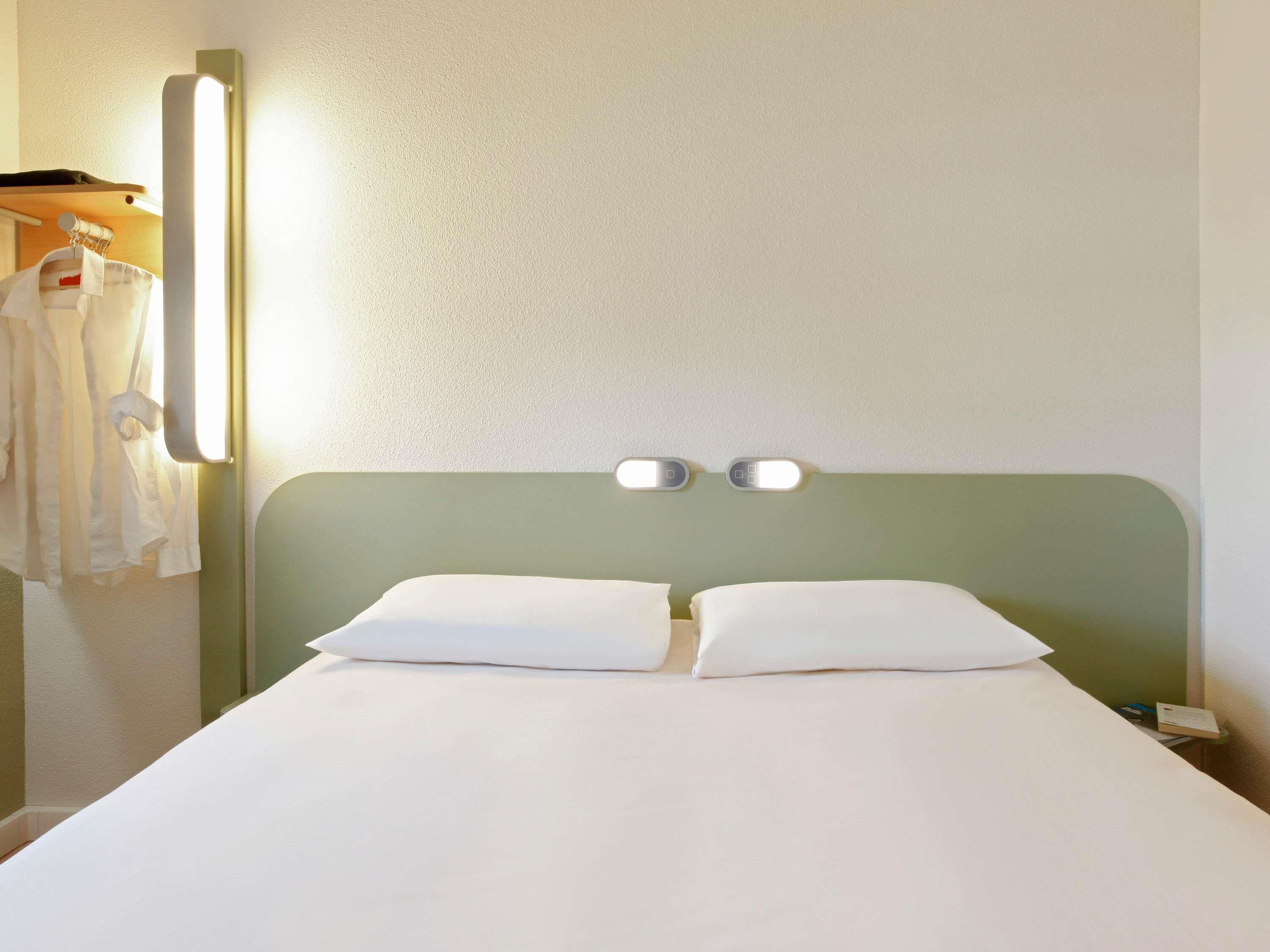 A hotel room with a bed, two bedside tables, a wall-mounted light, and a coat rack with hanging clothes.