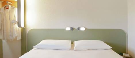 A hotel room with a bed, two bedside tables, a wall-mounted light, and a coat rack with hanging clothes.