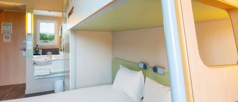 Una cama litera con una ventana pequeña, un lavamanos y un espejo en una habitación con puerta de madera y vista a árboles.