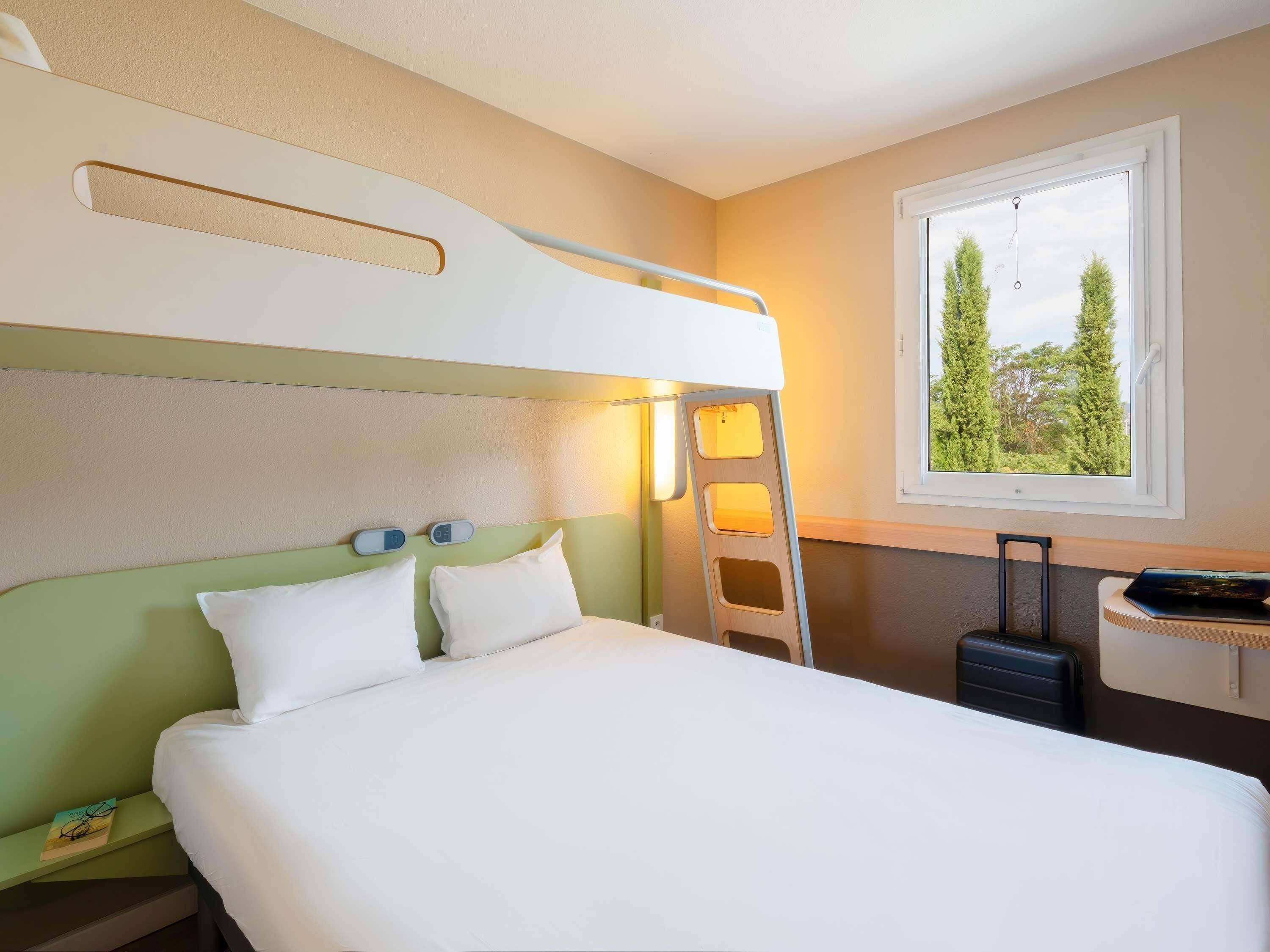 Un lit superposé avec une échelle, un bureau avec une valise et une fenêtre donnant sur des arbres.