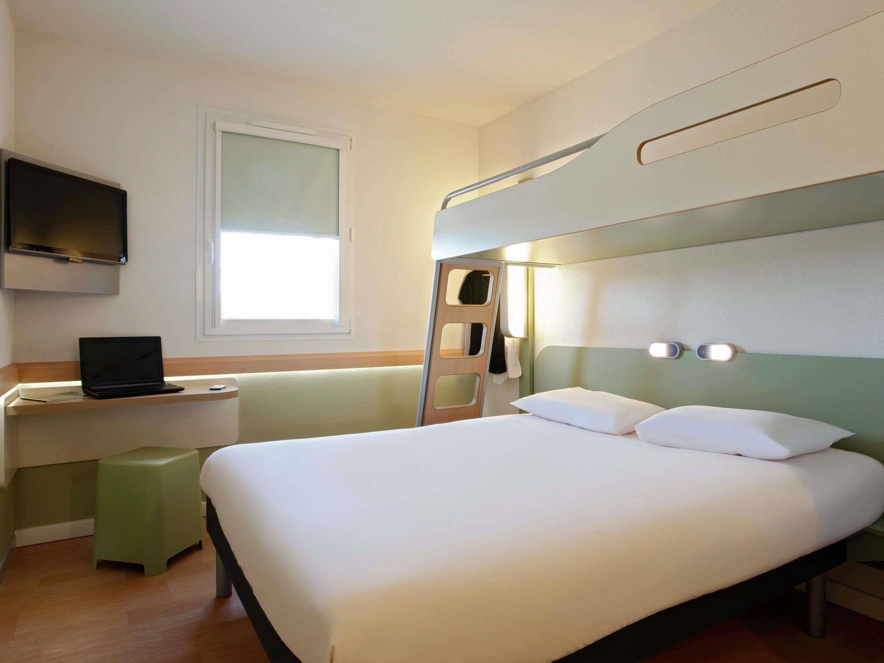 A hotel room with a bunk bed, a desk with a laptop, a television mounted on the wall, and a window providing natural light.