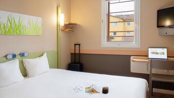 Ett hotellrum med en säng, ett skrivbord med en bärbar dator, en tv och ett fönster med utsikt över byggnader.