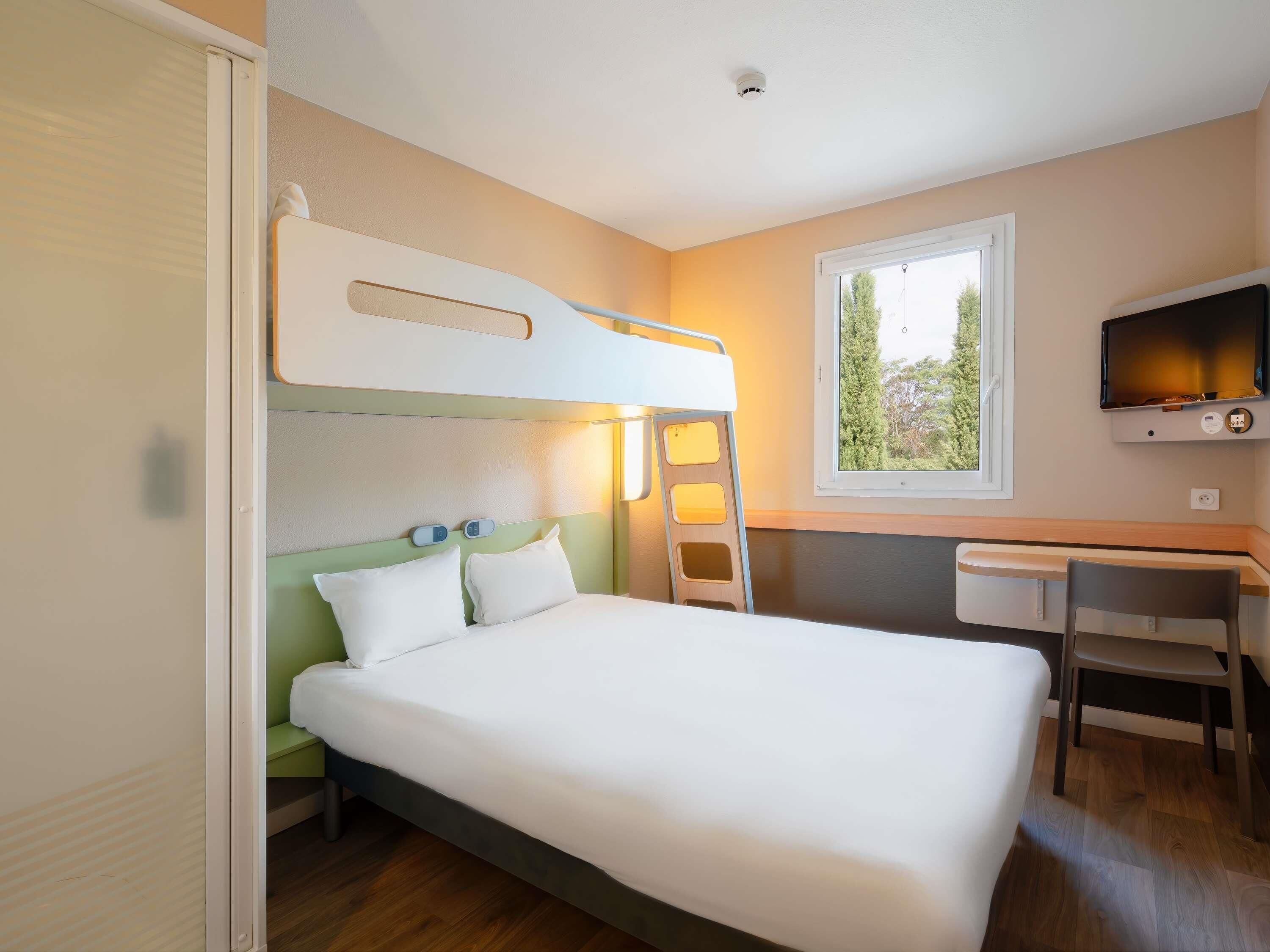 Une chambre avec un lit superposé, un bureau avec une chaise, une télévision et une fenêtre donnant sur des arbres.