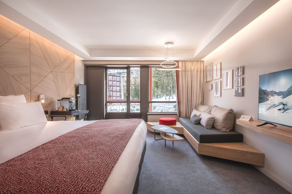 A modern hotel room with a large bed, a seating area with a red ottoman, a flat-screen TV, and a view of a snowy mountain.