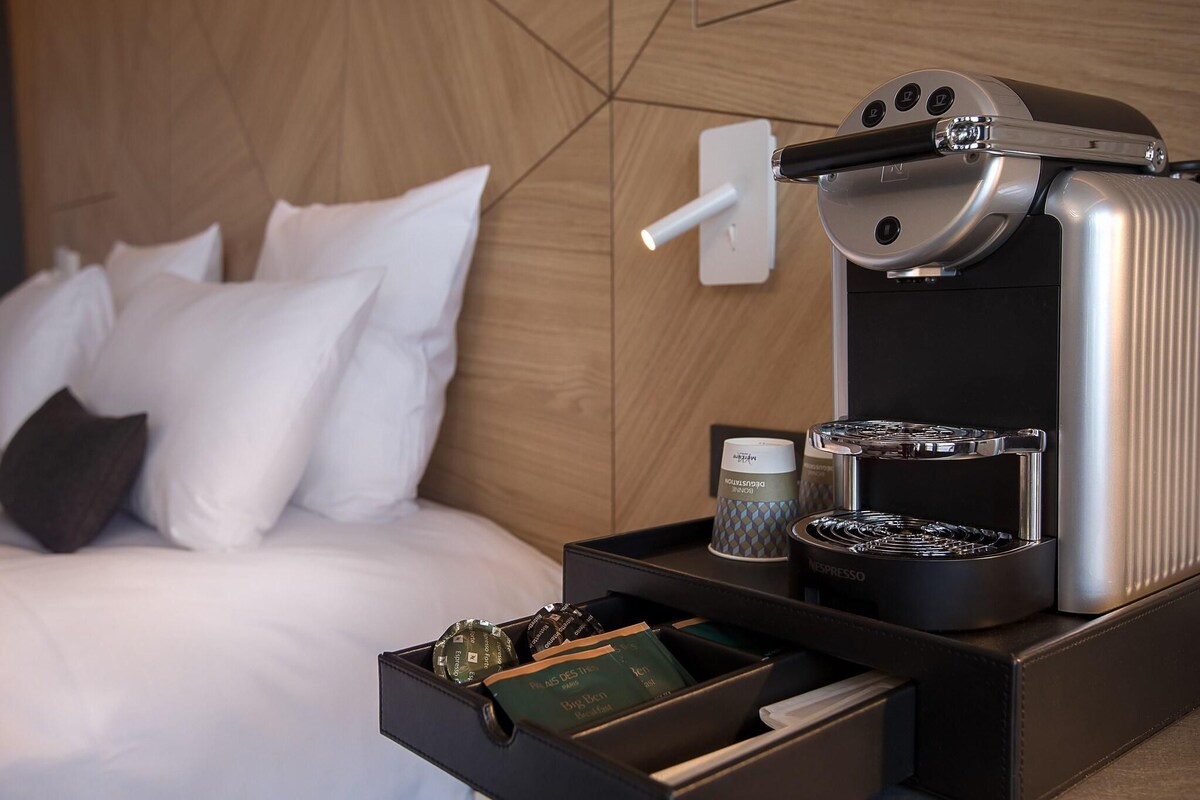 A hotel room with a bed, a wall-mounted espresso machine, a tray with coffee pods, a cup, and a box of amenities.