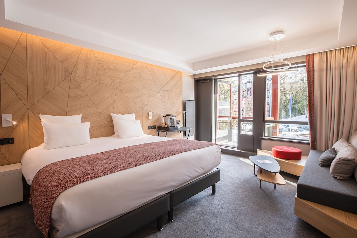 A modern hotel room with a large bed, a sofa, a small table, and a view of the city through large windows.