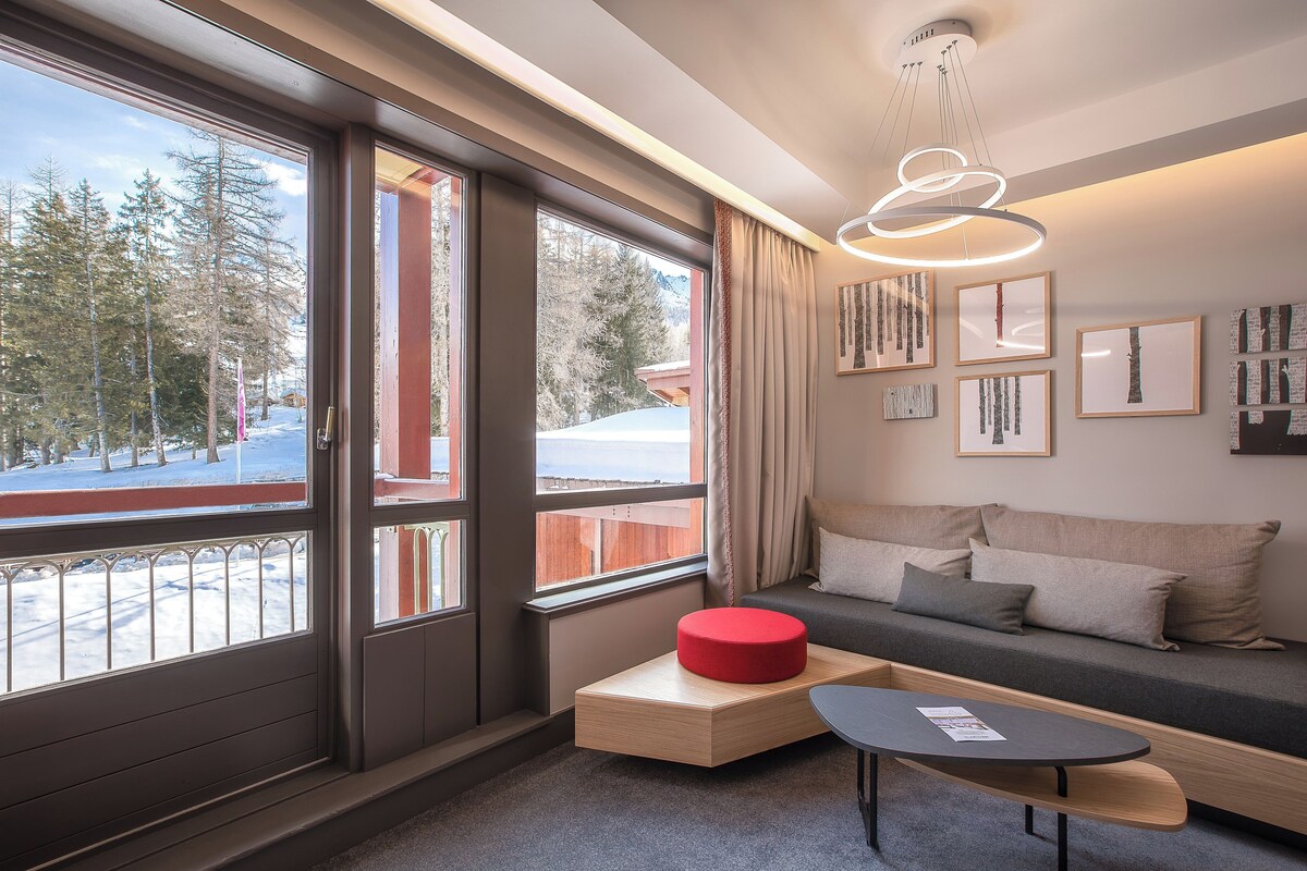 A modern interior with large windows, a sofa, a red ottoman, and a small table.