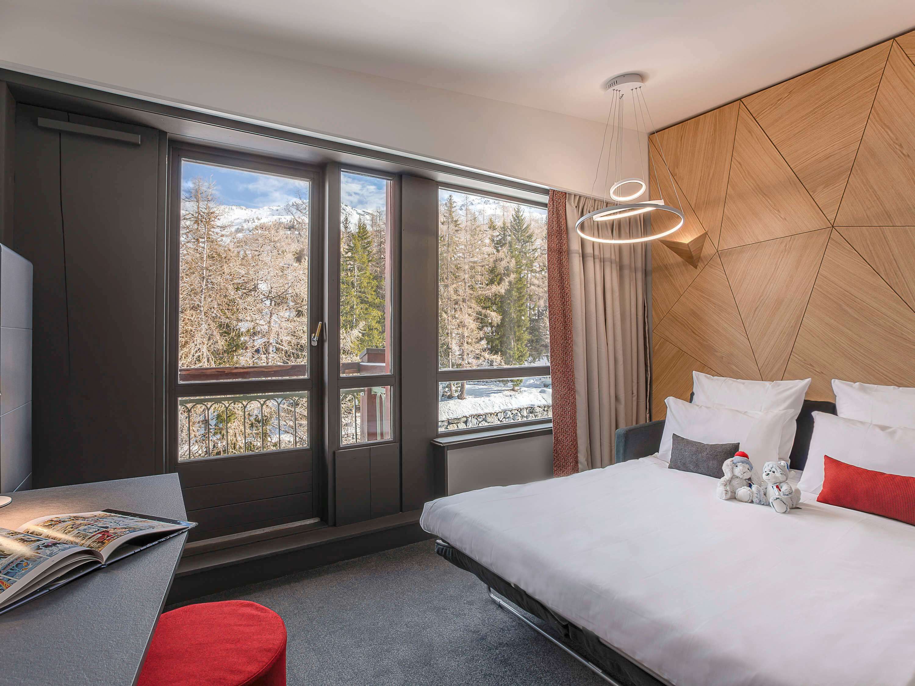 Een moderne hotelkamer met een groot bed, een bureau en uitzicht op een besneeuwd berglandschap door grote ramen.