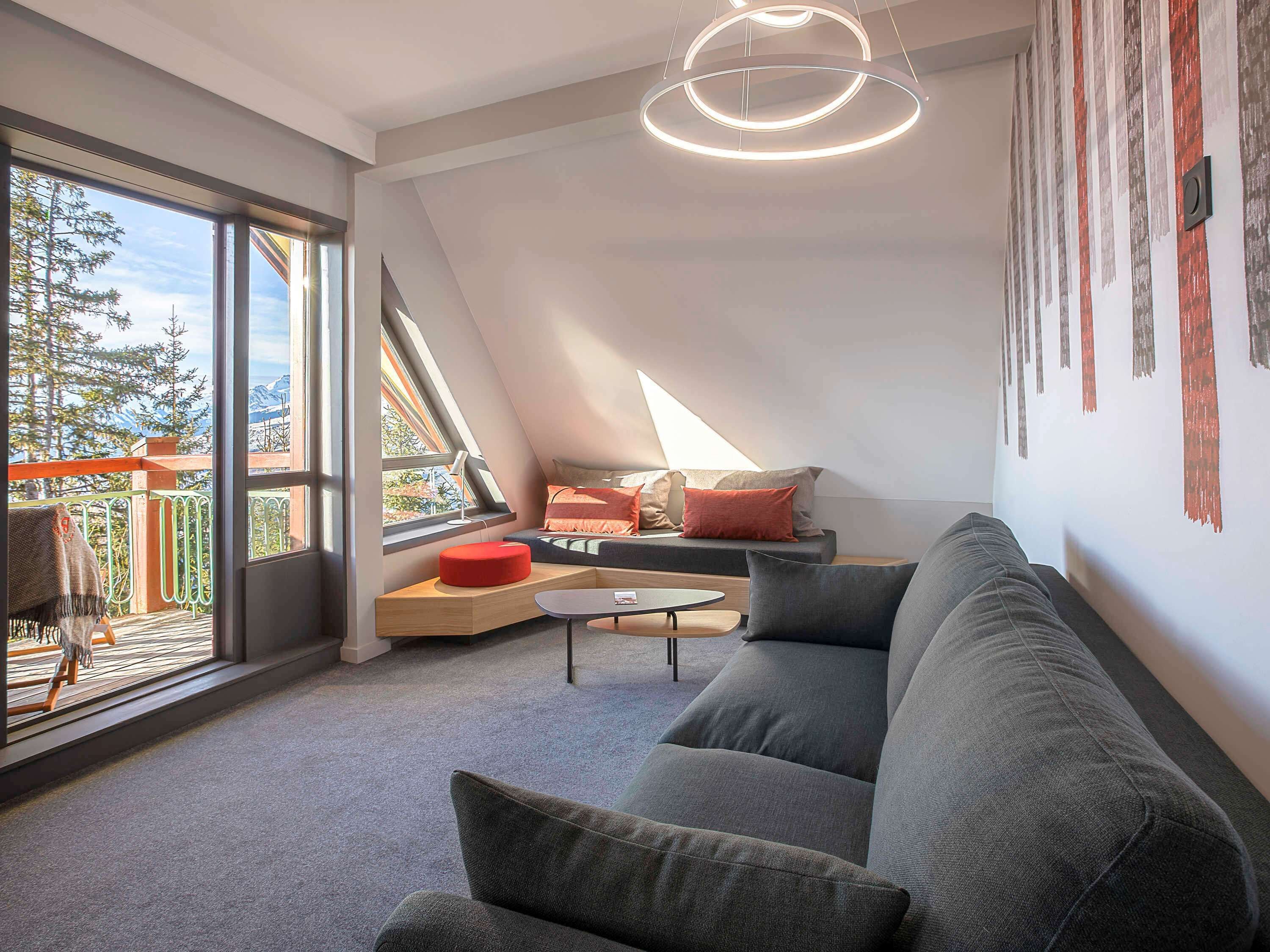 A modern loft bedroom with a sloped ceiling, large windows, a grey sofa, a red ottoman, and a circular light fixture.