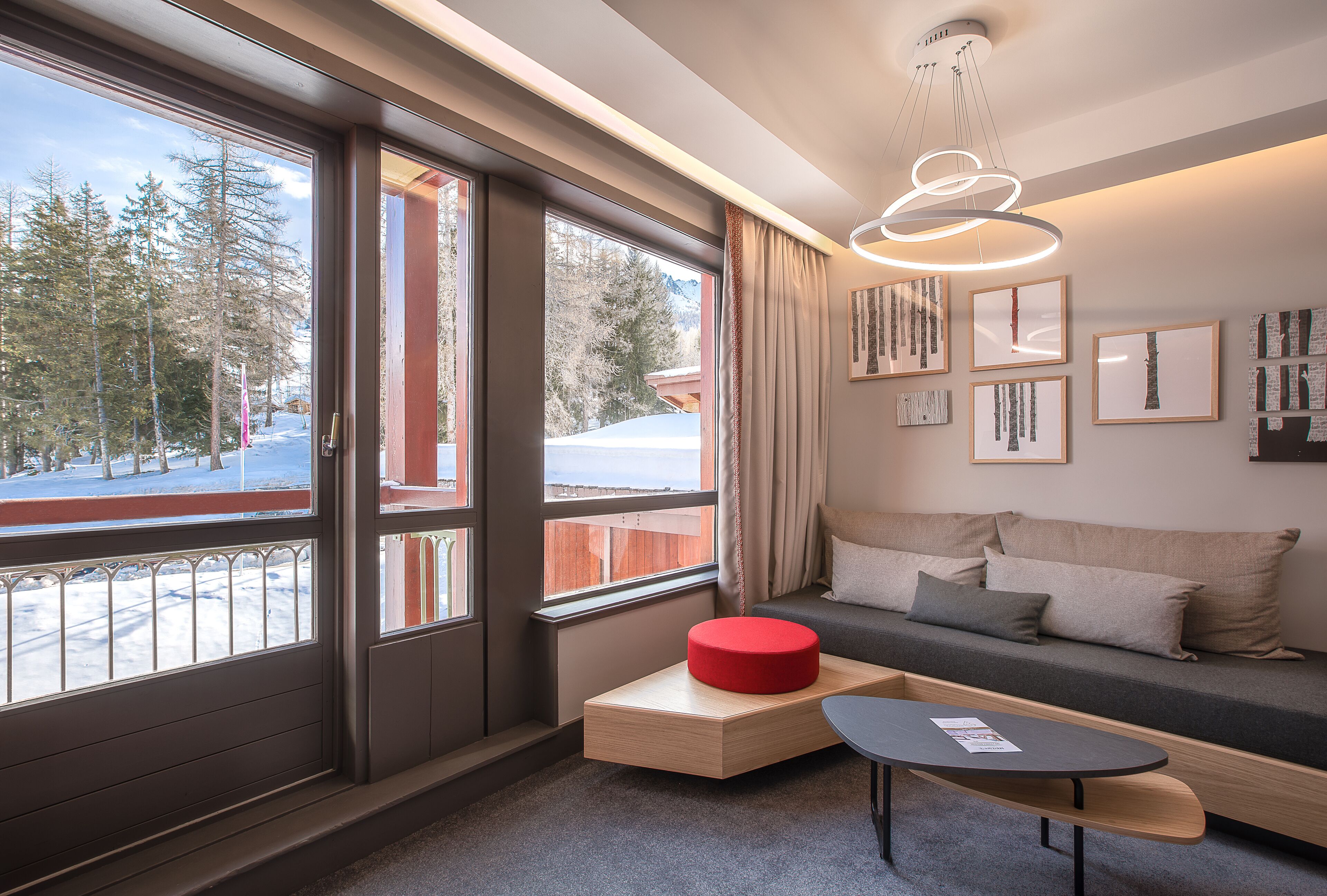 A modern room with large windows, a sofa, a small table, and a red ottoman.