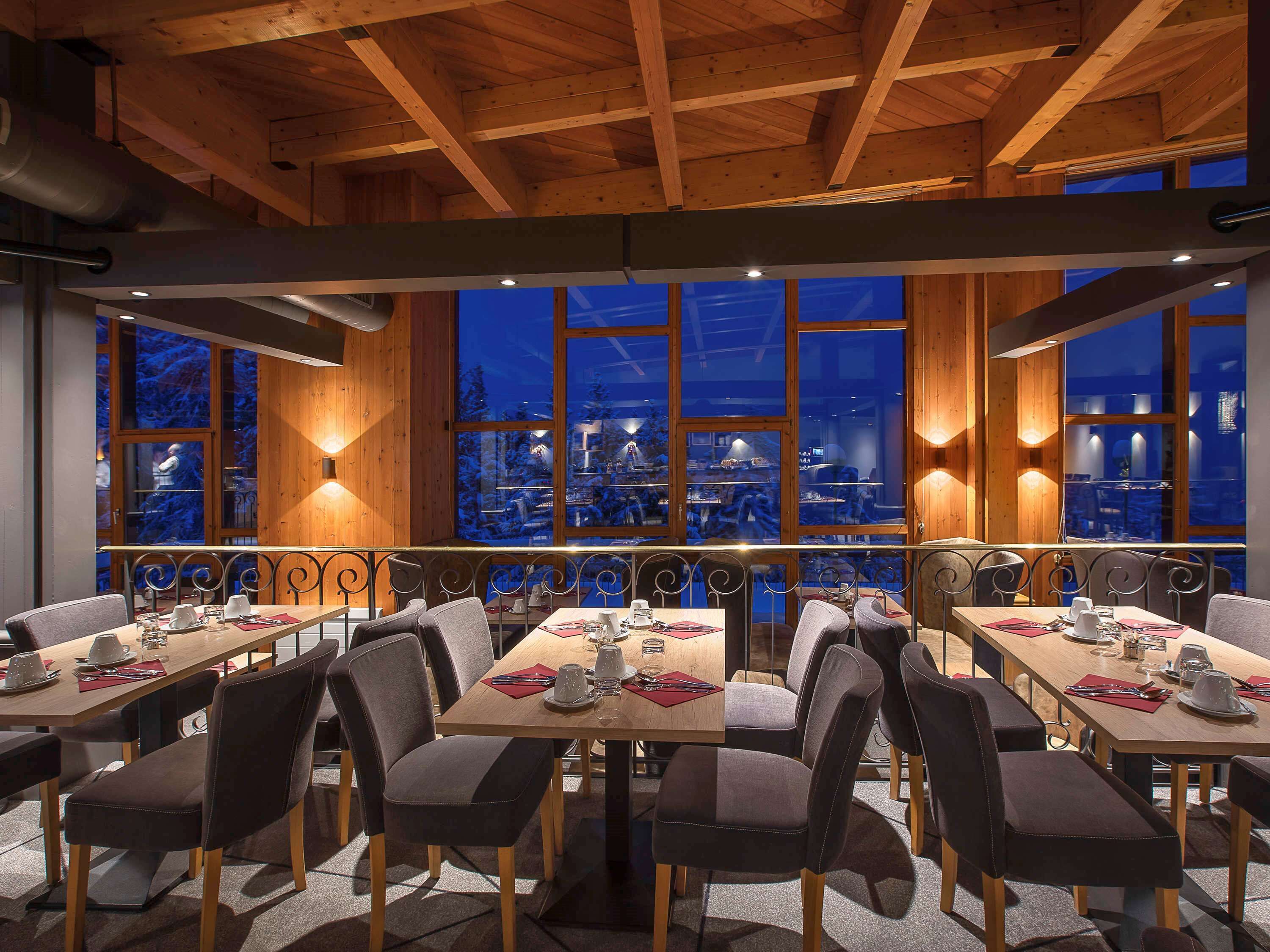 Restaurante com vigas de madeira e janelas amplas com vista para uma paisagem coberta de neve. Mesas montadas com talheres e copos.