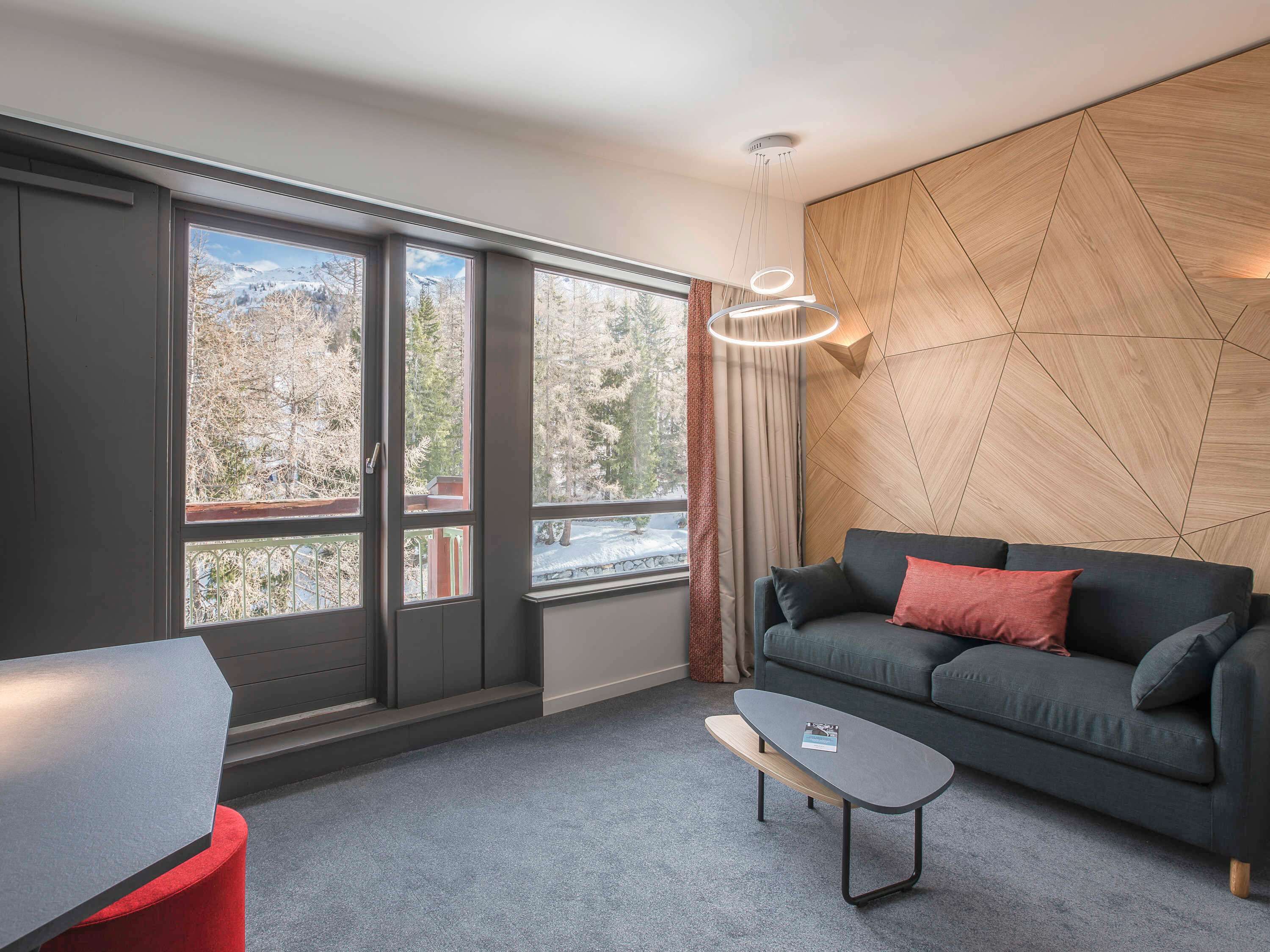 Un salon moderne avec un canapé gris foncé, une petite table basse et une vue sur un paysage enneigé, visible à travers de grandes fenêtres.