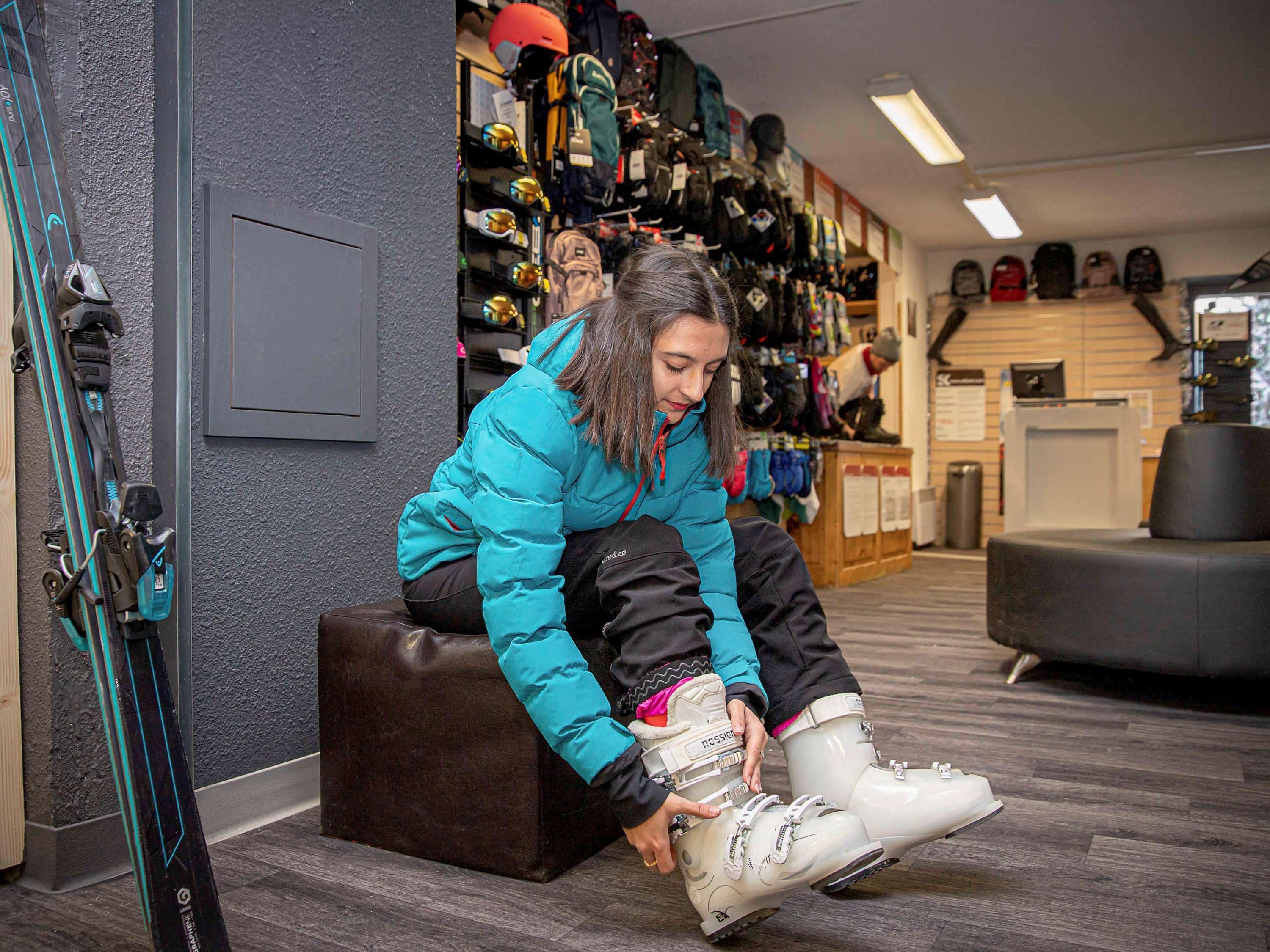A person sitting on a bench, tying ski boots in a room with skis and ski equipment on the wall.