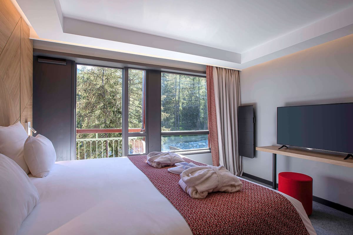 A hotel room with a large bed, a flat-screen TV on a stand, a balcony with a railing, and a view of trees and a body of water.