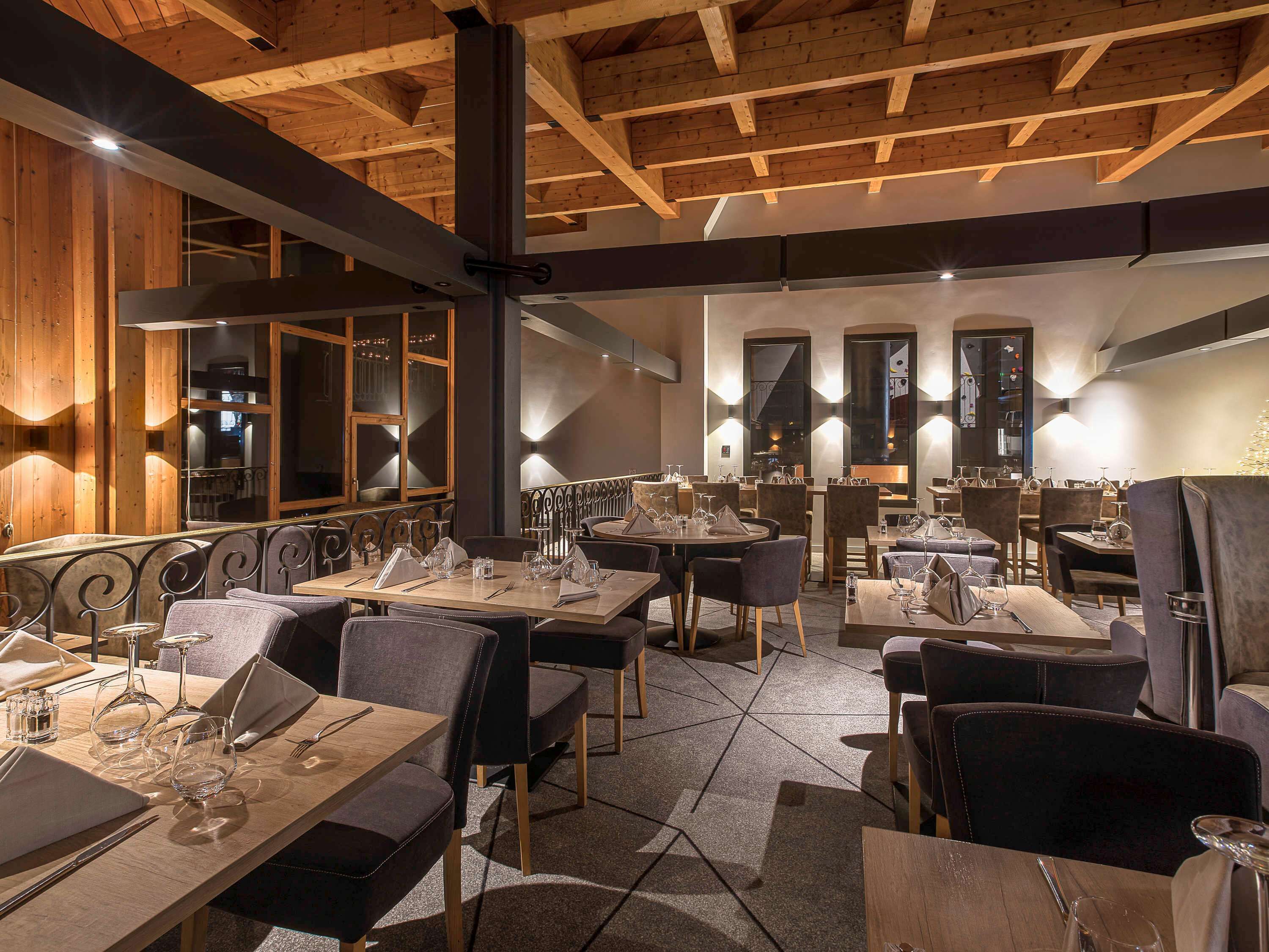 Un restaurante con vigas de madera en el techo, mesas dispuestas con copas de vino y cubiertos, y vistas a la cocina a través de un amplio ventanal.