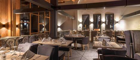 Un restaurante con vigas de madera en el techo, mesas dispuestas con copas de vino y cubiertos, y vistas a la cocina a través de un amplio ventanal.