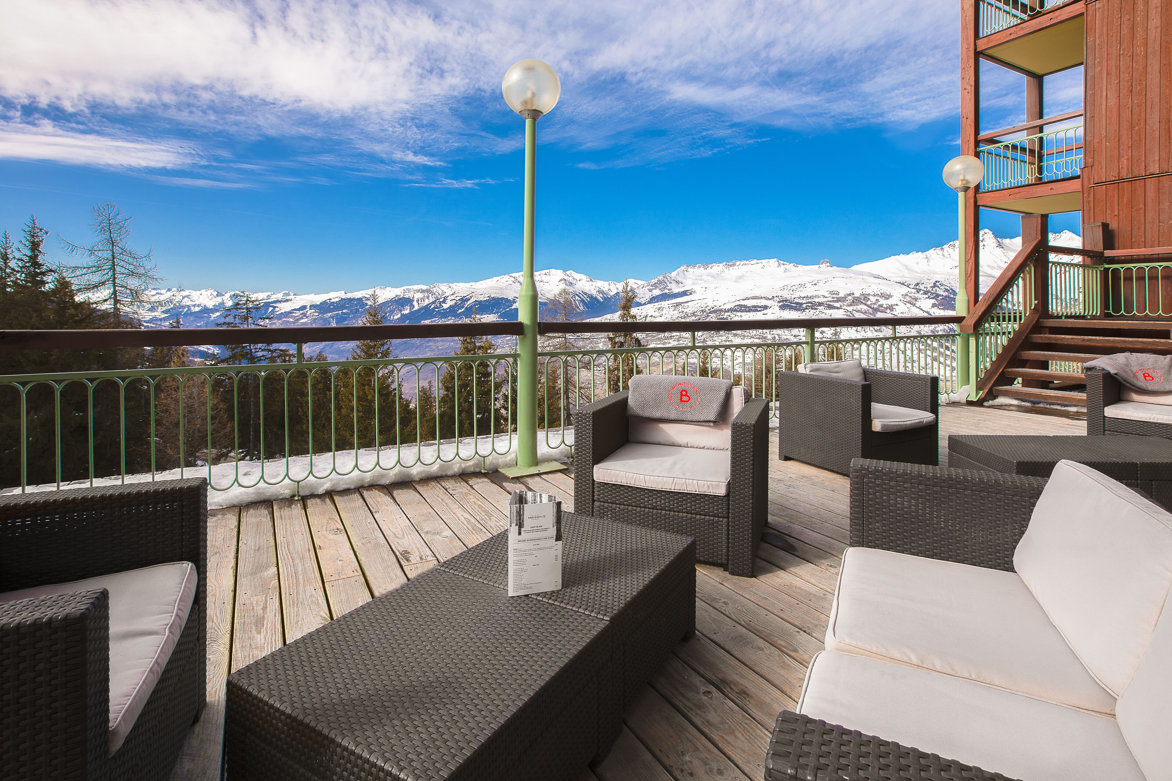 Une terrasse dotée de meubles en osier, d’un escalier en bois et offrant une vue sur des montagnes enneigées.