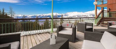 Une terrasse dotée de meubles en osier, d’un escalier en bois et offrant une vue sur des montagnes enneigées.