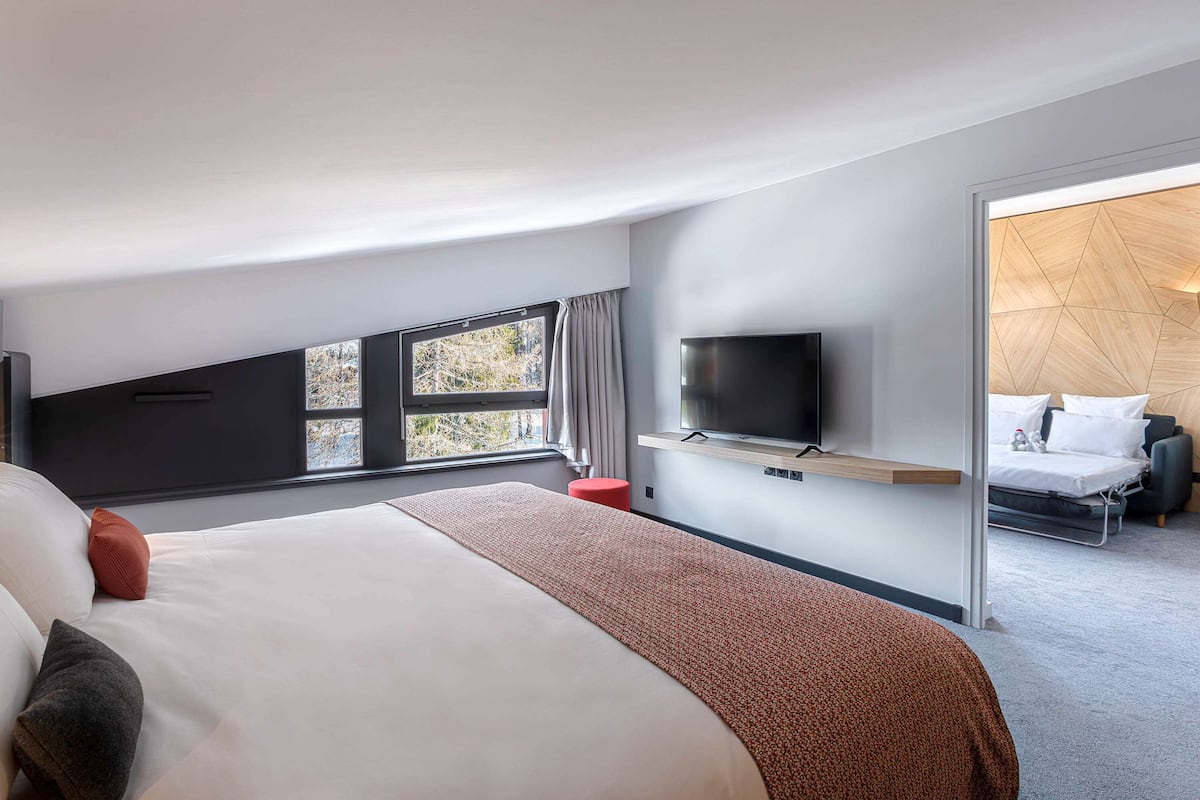 A modern hotel room with a large bed, a flat-screen TV mounted on the wall, a window with a view of trees, and a wooden accent wall.