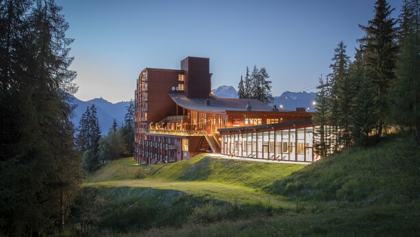 Un edificio moderno con fachada de vidrio, rodeado de árboles y montañas.