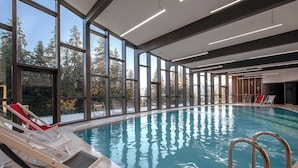 Una piscina interior con vistas despejadas a árboles a través de grandes ventanales, sillones de descanso y un diseño moderno de techo.