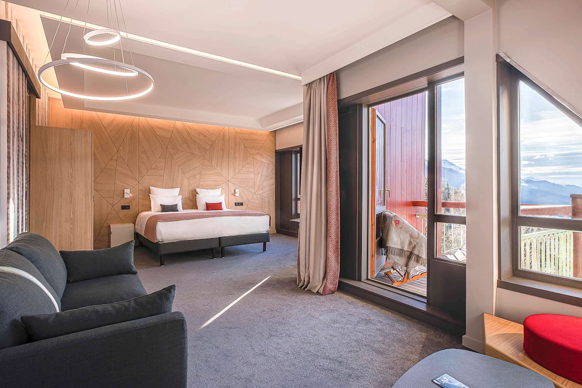 A modern hotel room with a large bed, a sofa, a small table, and a view of mountains through the window.
