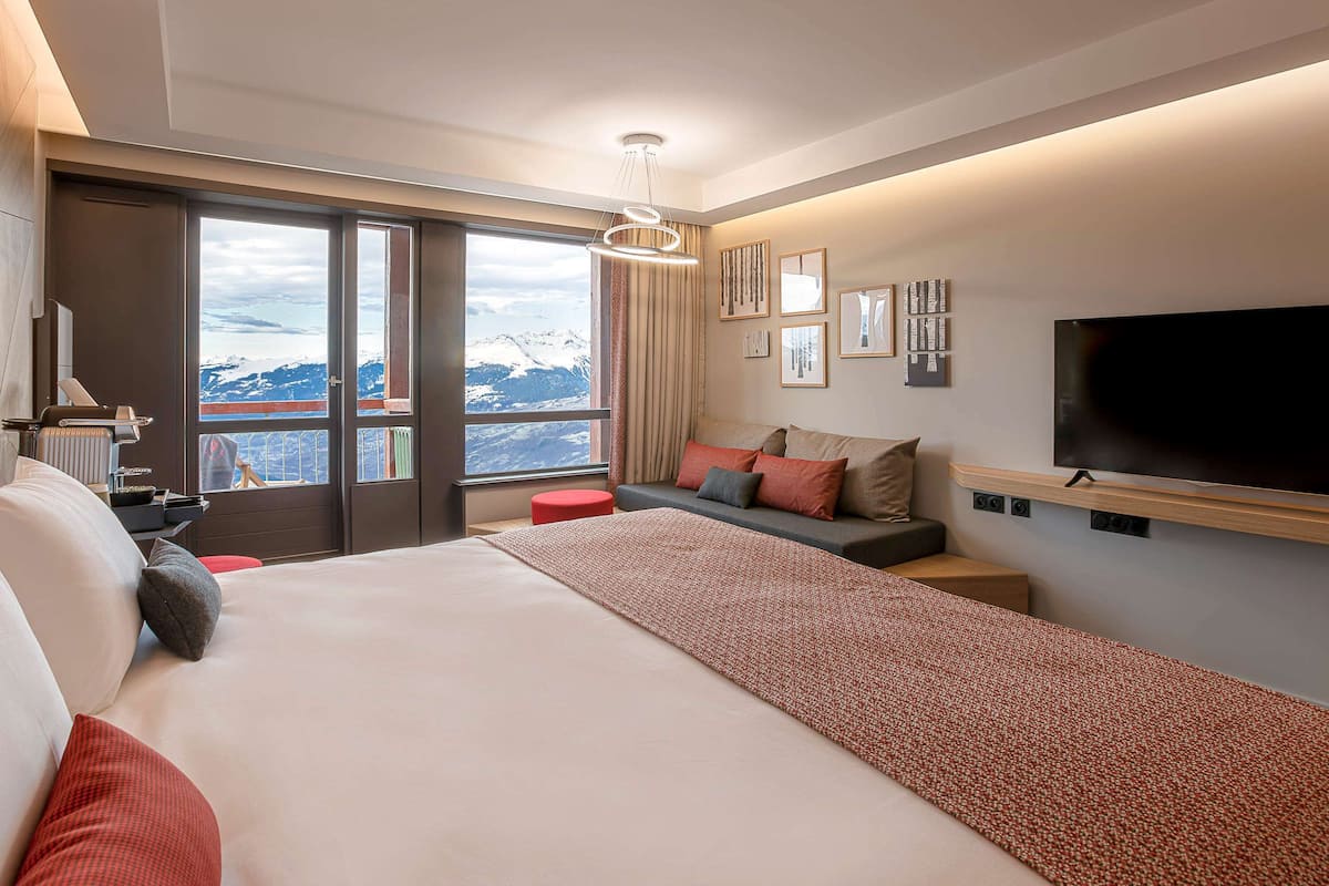 A modern hotel room with a large bed, a flat-screen TV, and a view of mountains through large windows.