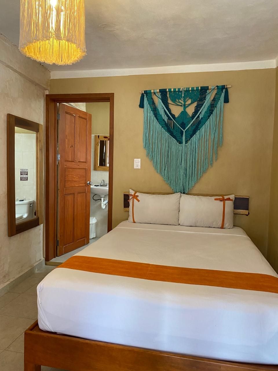 A bedroom with a bed, a wooden headboard, a wall-mounted macrame piece, a mirror, and a door leading to a bathroom.