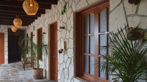 Un pasillo con paredes de piedra, puertas y ventanas de madera, plantas en macetas y lámparas de mimbre colgantes.