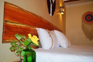 Un letto con biancheria bianca e un poggiatesta in legno, una bottiglia verde con un fiore giallo e un elemento decorativo a parete.