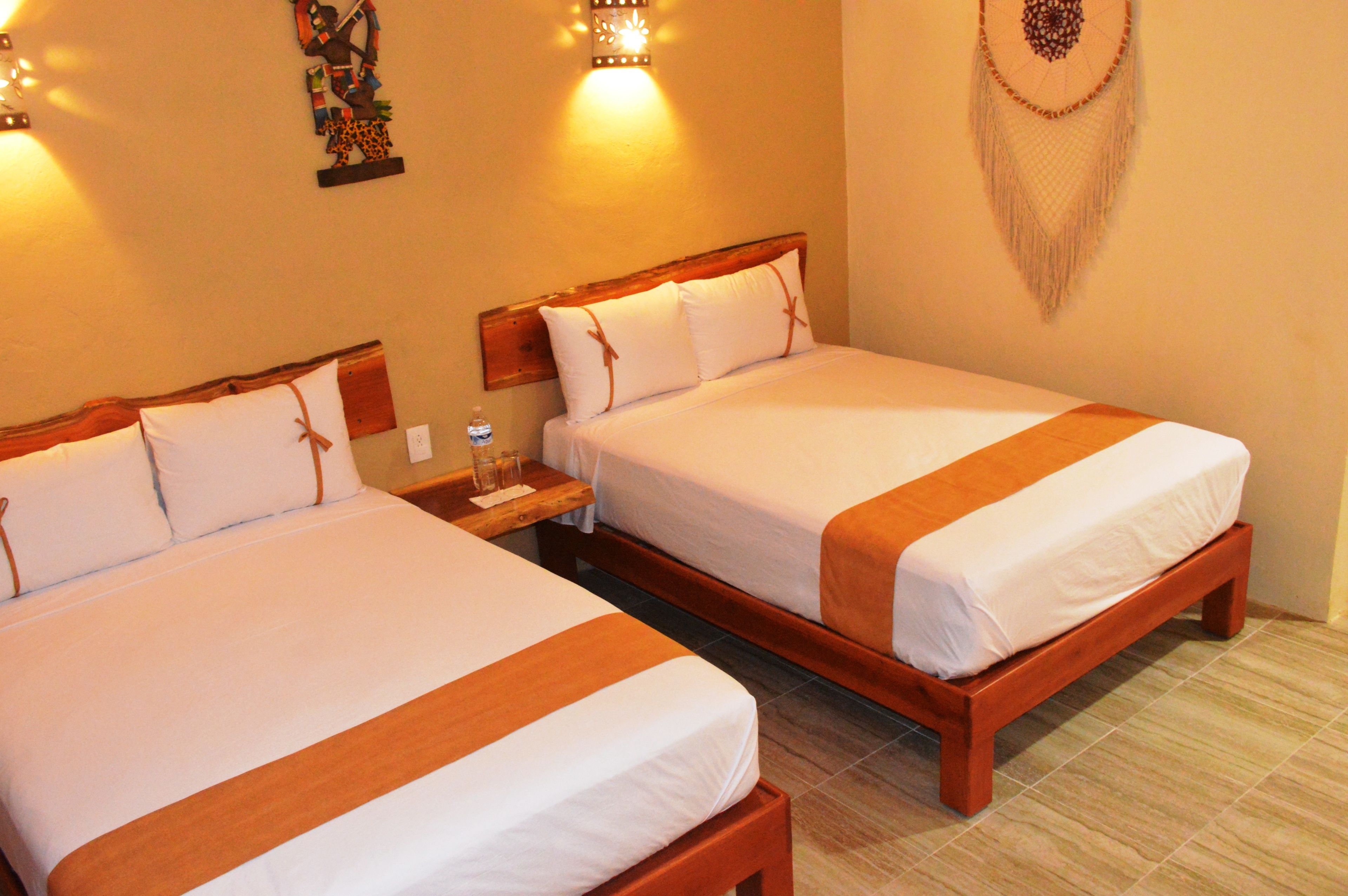Dos camas matrimoniales con cabeceros de madera y ropa de cama blanca, una mesita de noche con un vaso de agua, y detalles decorativos en la pared, incluyendo un atrapasueños y una escultura de jirafa.