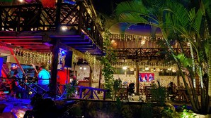 Un restaurante al aire libre con zona de estar cubierta, paredes de piedra y luces colgantes.