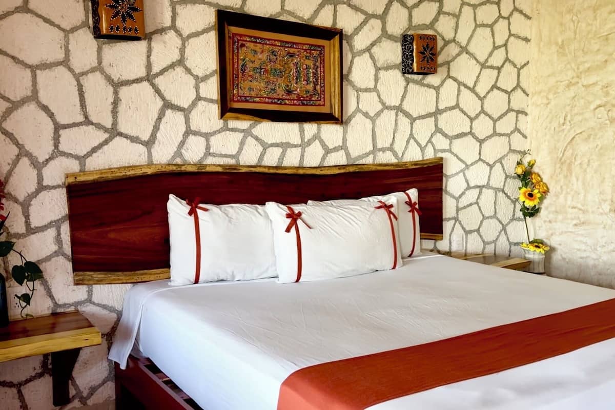 A bed with white linens and red accents, a wooden headboard, and a stone wall.