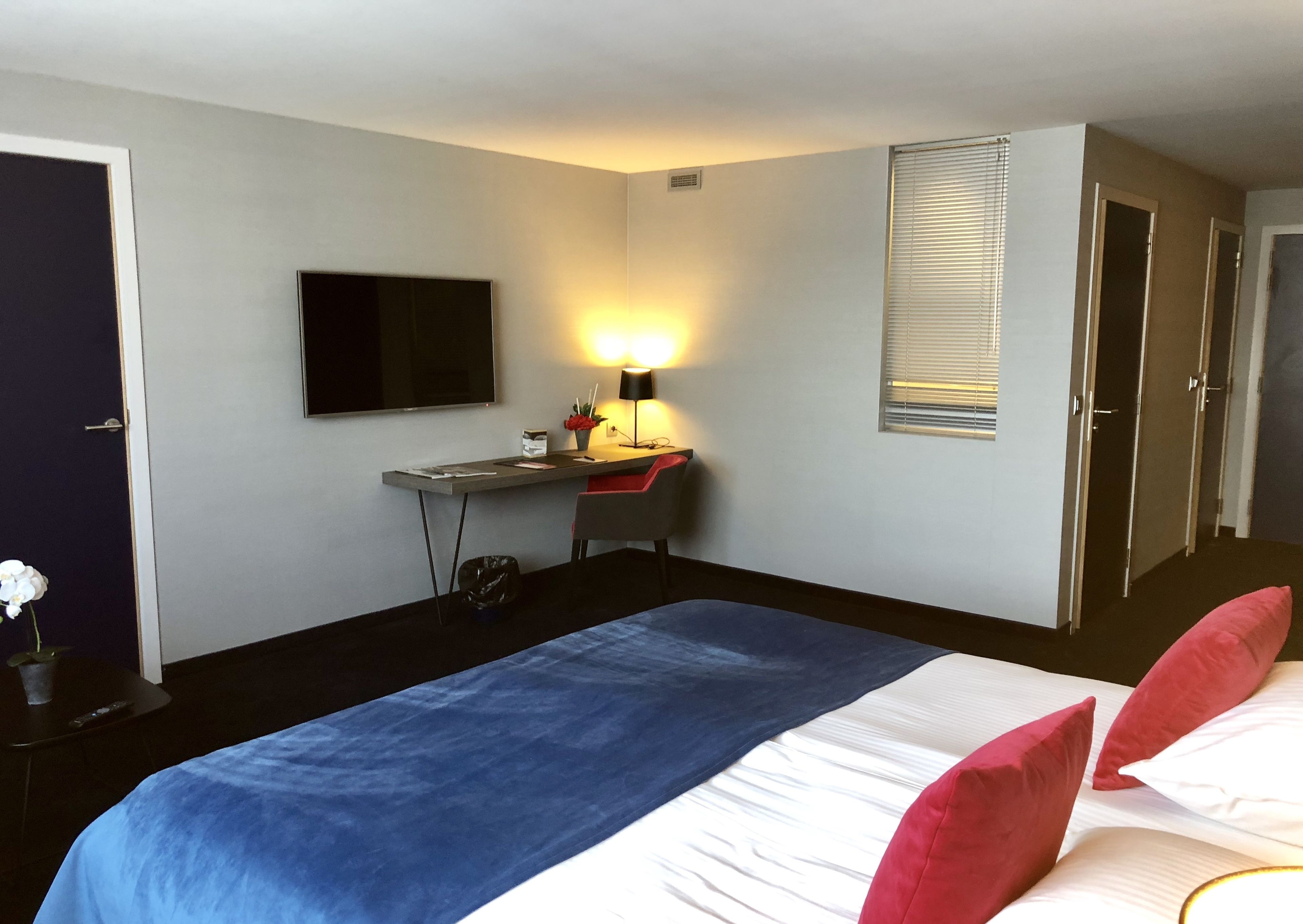 Una habitación de hotel con cama, escritorio, silla, televisor y ventana con persianas.