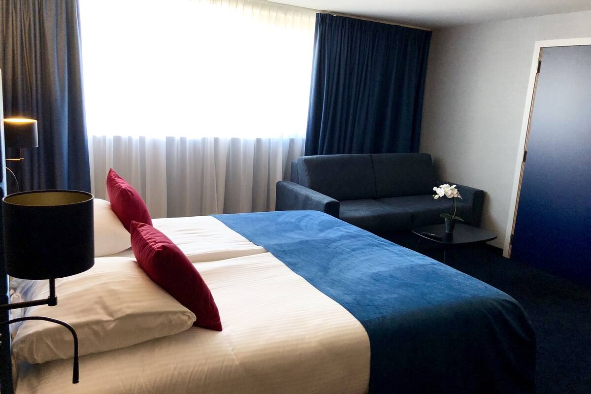 Une chambre d’hôtel avec un lit, un canapé, une petite table avec une plante et une fenêtre avec des rideaux.