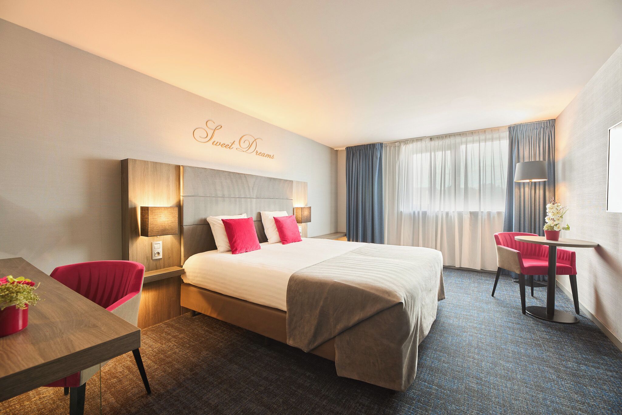 A hotel room with a bed, a desk, a chair, a lamp, and a window with curtains.