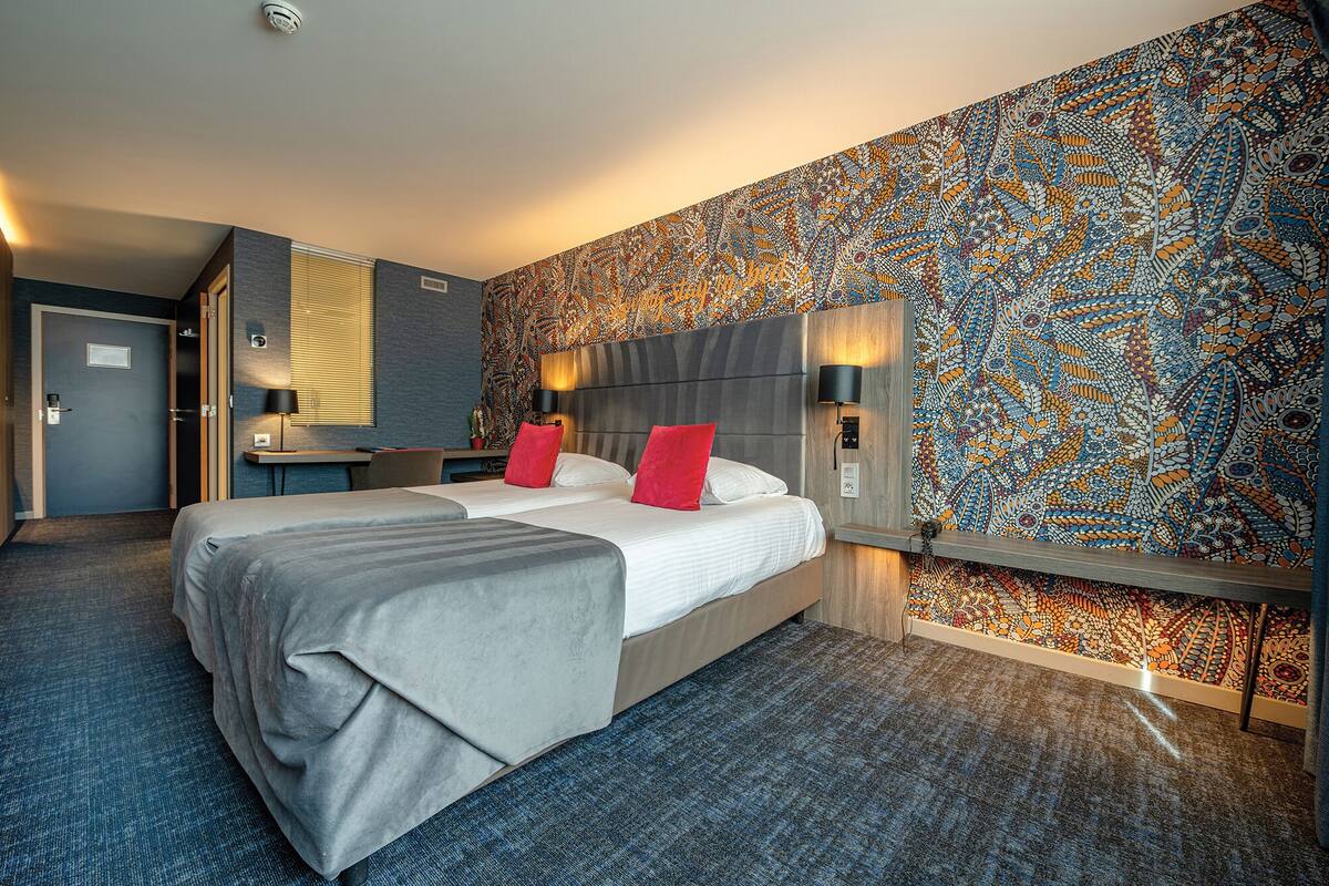 Une chambre d’hôtel avec un grand lit, deux tables de chevet, une petite table d’appoint, un téléviseur fixé au mur et un papier peint à motifs.