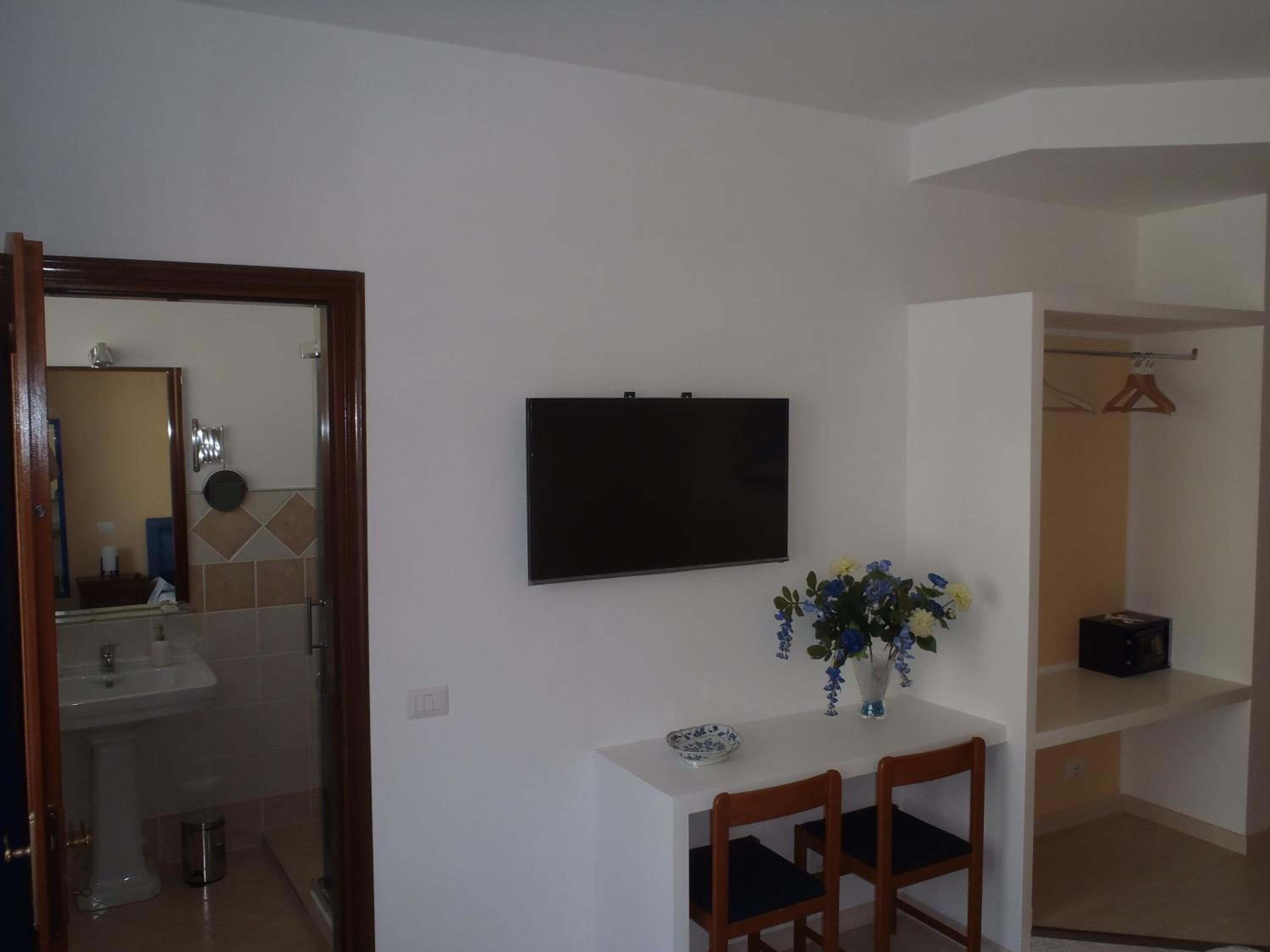 Una habitación pequeña con una mesa blanca, dos sillas, un televisor montado en la pared, un florero con flores y un espejo que refleja un baño.