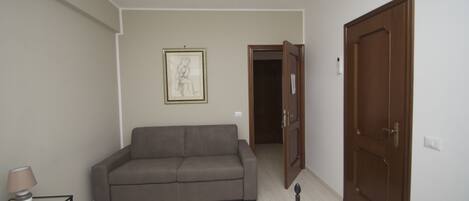 A room with a grey sofa, a small table with a lamp, a framed picture on the wall, and a wooden door.