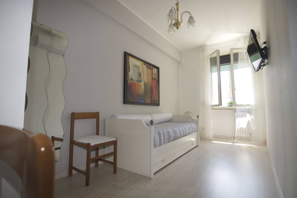 A room with a white bed, a wooden chair, a painting on the wall, and a window with a view.